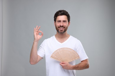 Photo of Happy man holding hand fan and showing ok gesture on light grey background