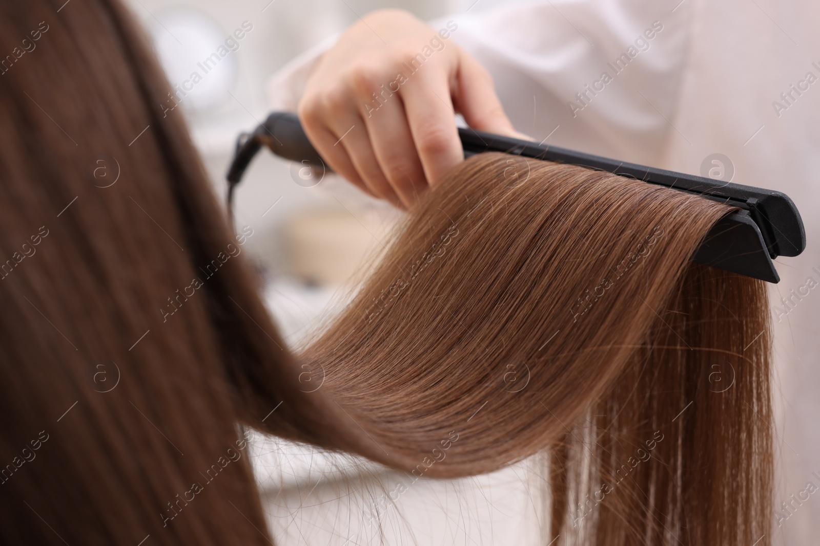 Photo of Hairdresser straightening woman's hair with flat iron indoors, closeup