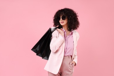 Happy young woman with shopping bags and stylish sunglasses on pink background