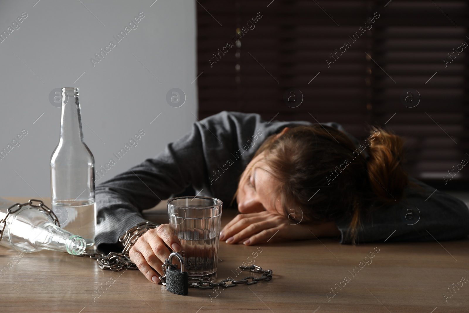 Photo of Alcohol addiction. Woman chained with glass of vodka sleeping on wooden table in room