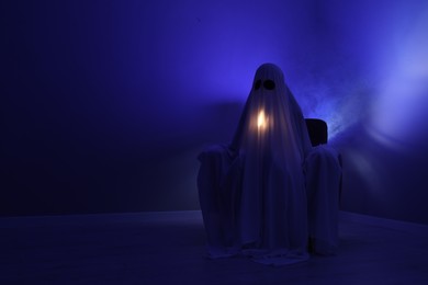 Photo of Creepy ghost. Woman covered with sheet sitting in armchair in blue light, space for text