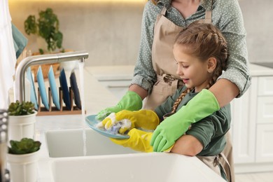 Photo of Mother and daughter wearing protective gloves washing plate above sink in kitchen