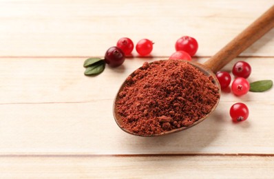 Photo of Dried cranberry powder in spoon, fresh berries and green leaves on light wooden table, closeup. Space for text