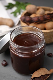 Photo of Jar of tasty tamarind sauce and fresh pods on grey wooden table