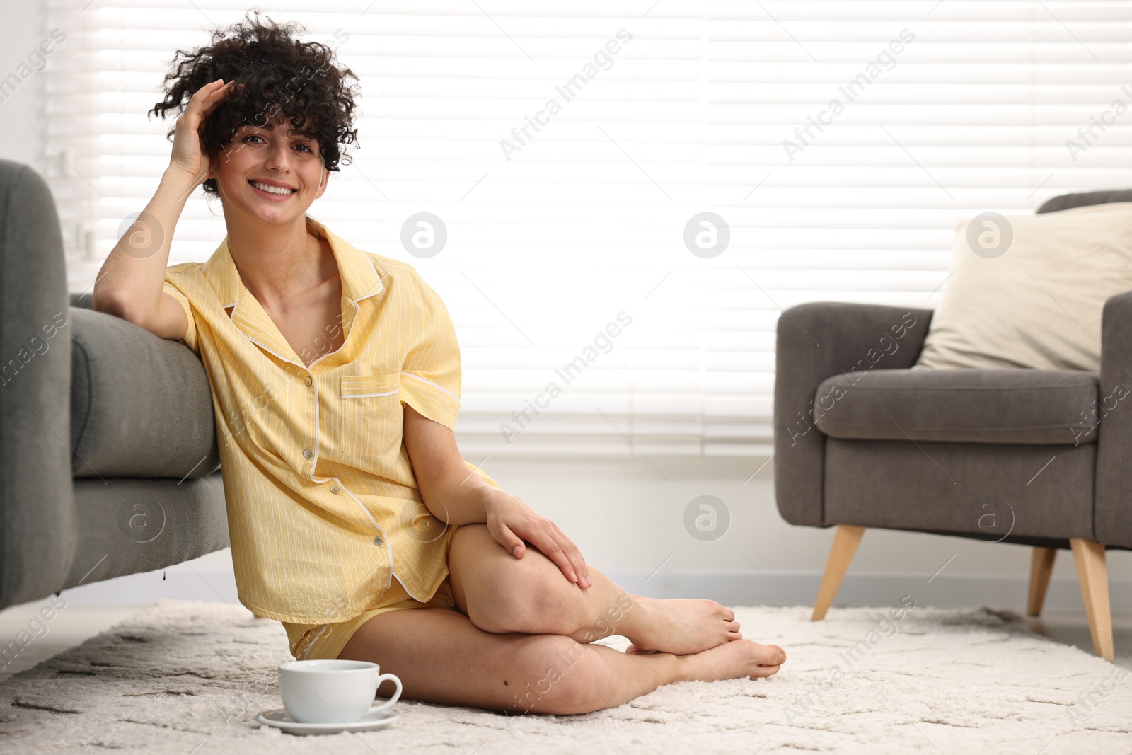 Photo of Beautiful young woman in stylish pyjama with cup of drink on floor at home