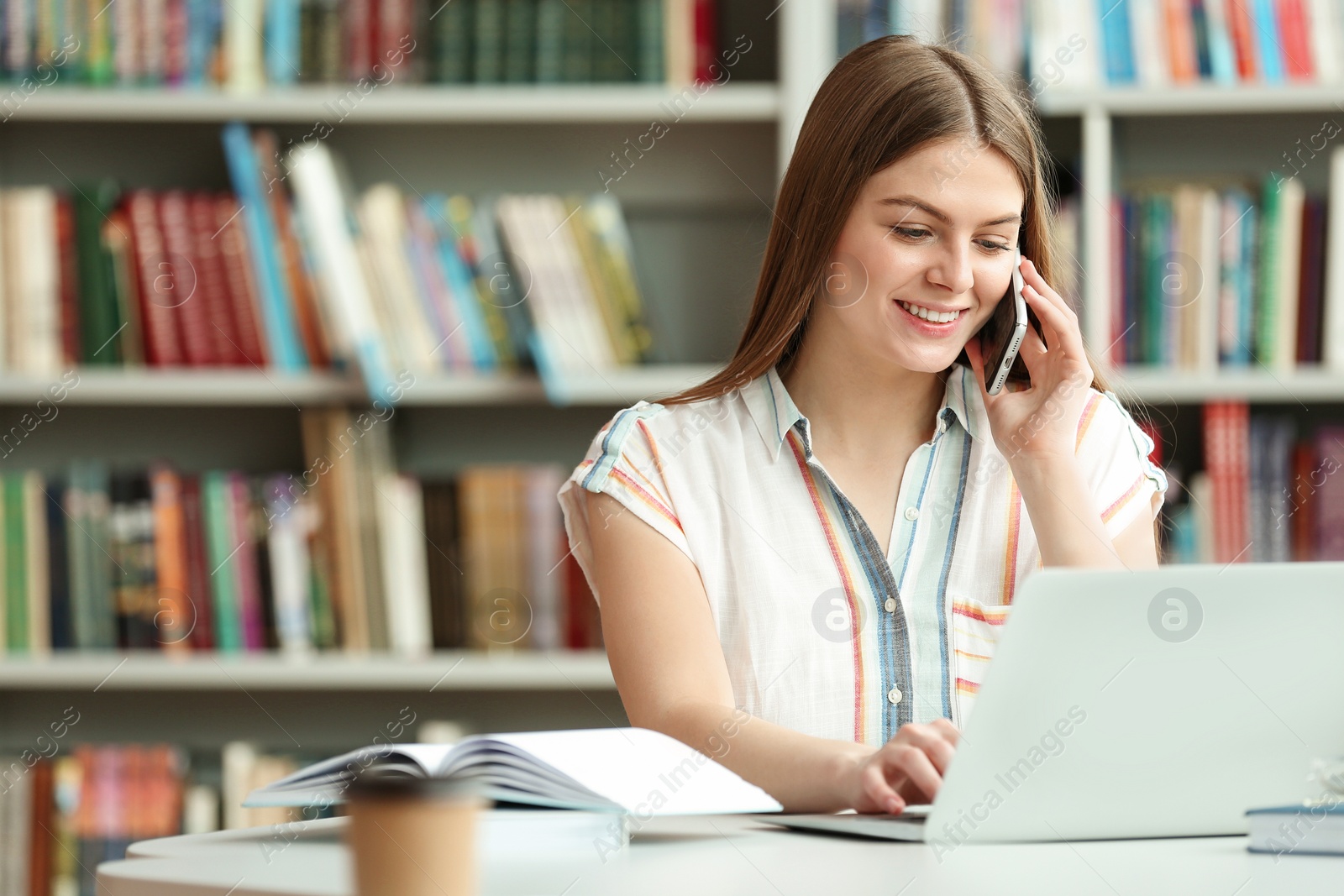 Photo of Young woman talking on phone and working with laptop at table in library. Space for text