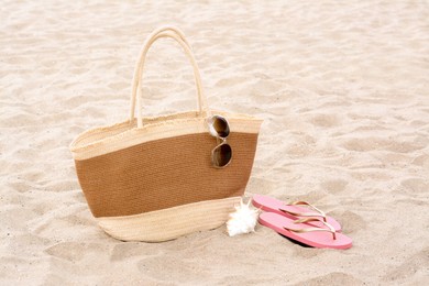 Photo of Stylish straw bag with sunglasses, flip flops and seashell on sand outdoors