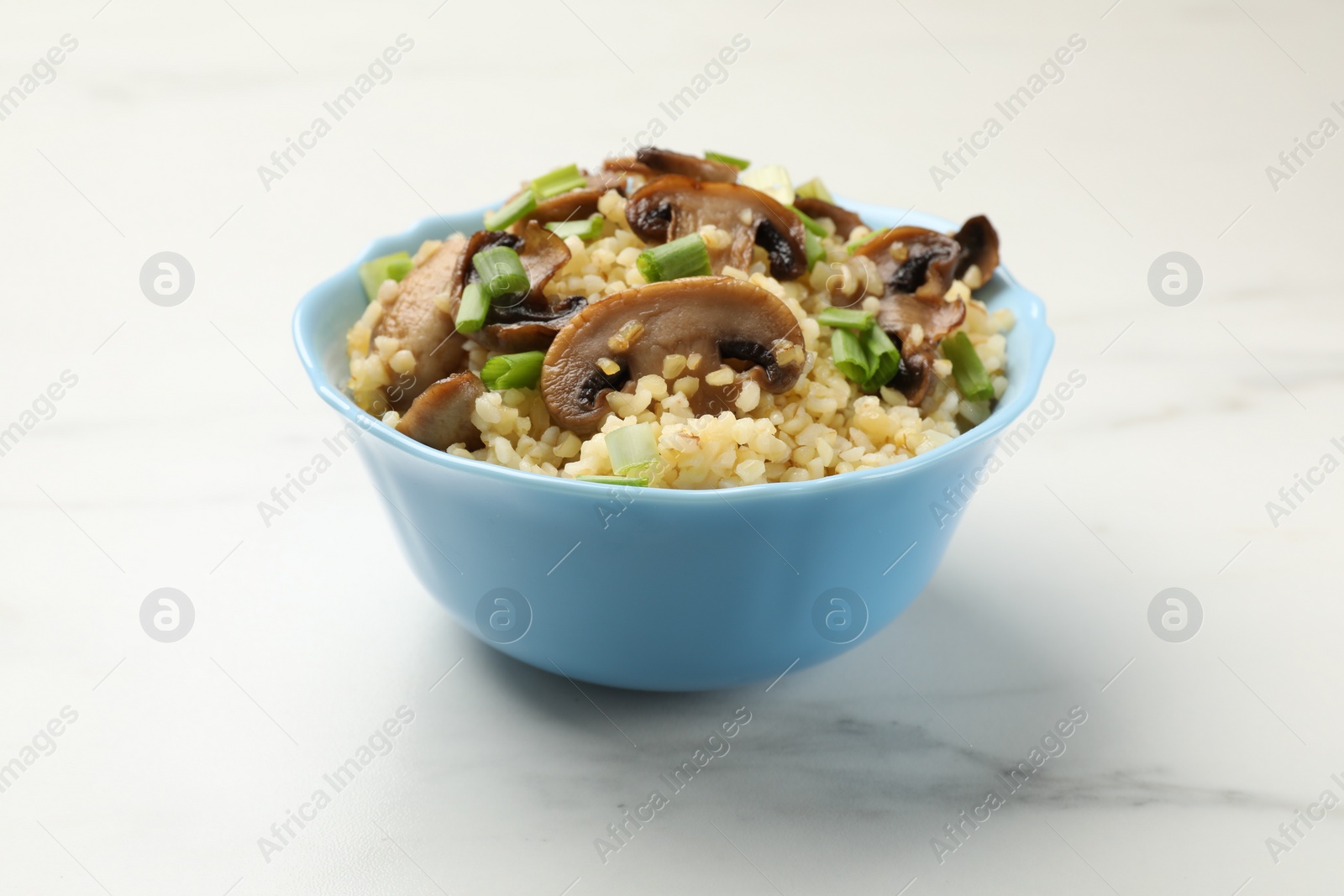 Photo of Delicious bulgur with mushrooms and green onion in bowl on white marble table, closeup