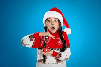 Photo of Girl in Santa hat with hourglass on light blue background. Christmas countdown