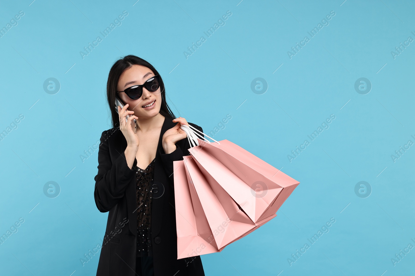 Photo of Smiling woman with shopping bags talking by smartphone on light blue background. Space for text