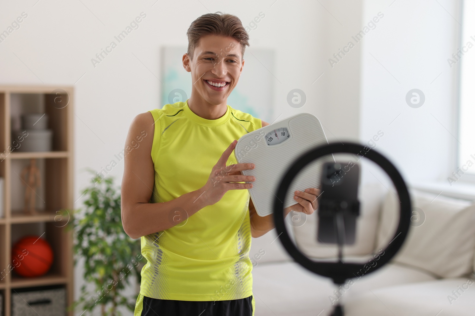 Photo of Smiling sports blogger holding floor scales while streaming online fitness lesson with phone at home
