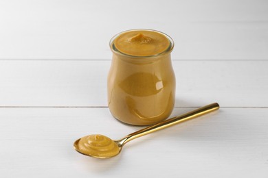 Photo of Spoon and glass jar of tasty mustard sauce on white wooden table