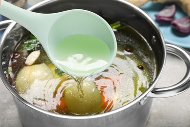 Ladle with tasty bouillon above pot at grey table, closeup