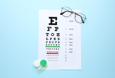 Case with contact lenses, tweezers, glasses and eye chart test on light blue background, flat lay