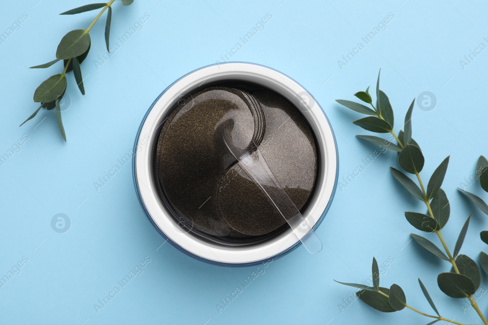 Photo of Under eye patches in jar with spatula and green twigs on light blue background, flat lay. Cosmetic product