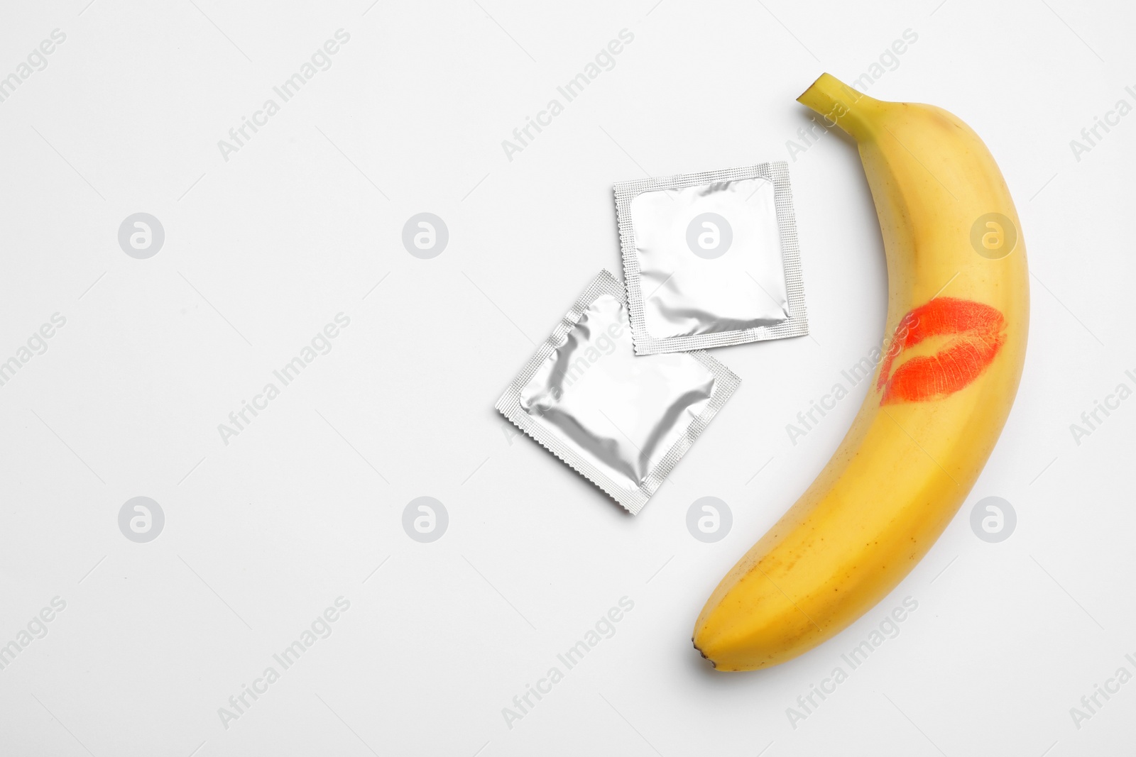 Photo of Condoms and banana with lipstick kiss mark on white background, top view. Safe sex