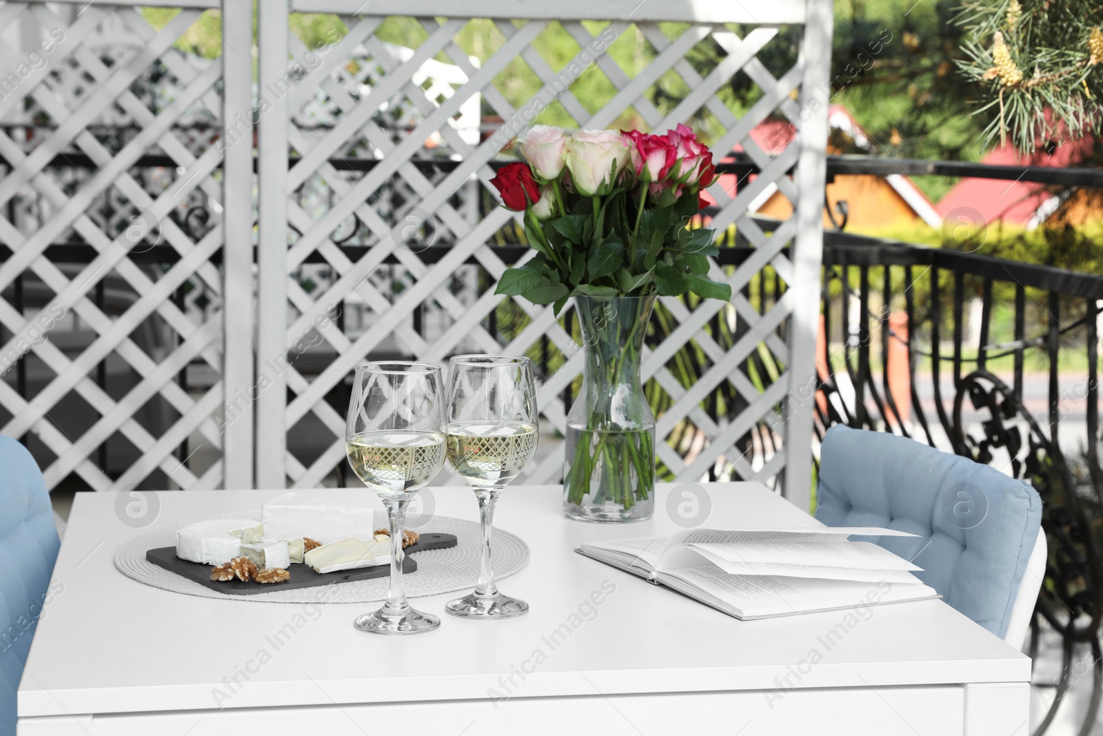 Photo of Vase with roses, open book, glasses of wine and snacks on white table at balcony