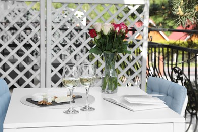 Photo of Vase with roses, open book, glasses of wine and snacks on white table at balcony