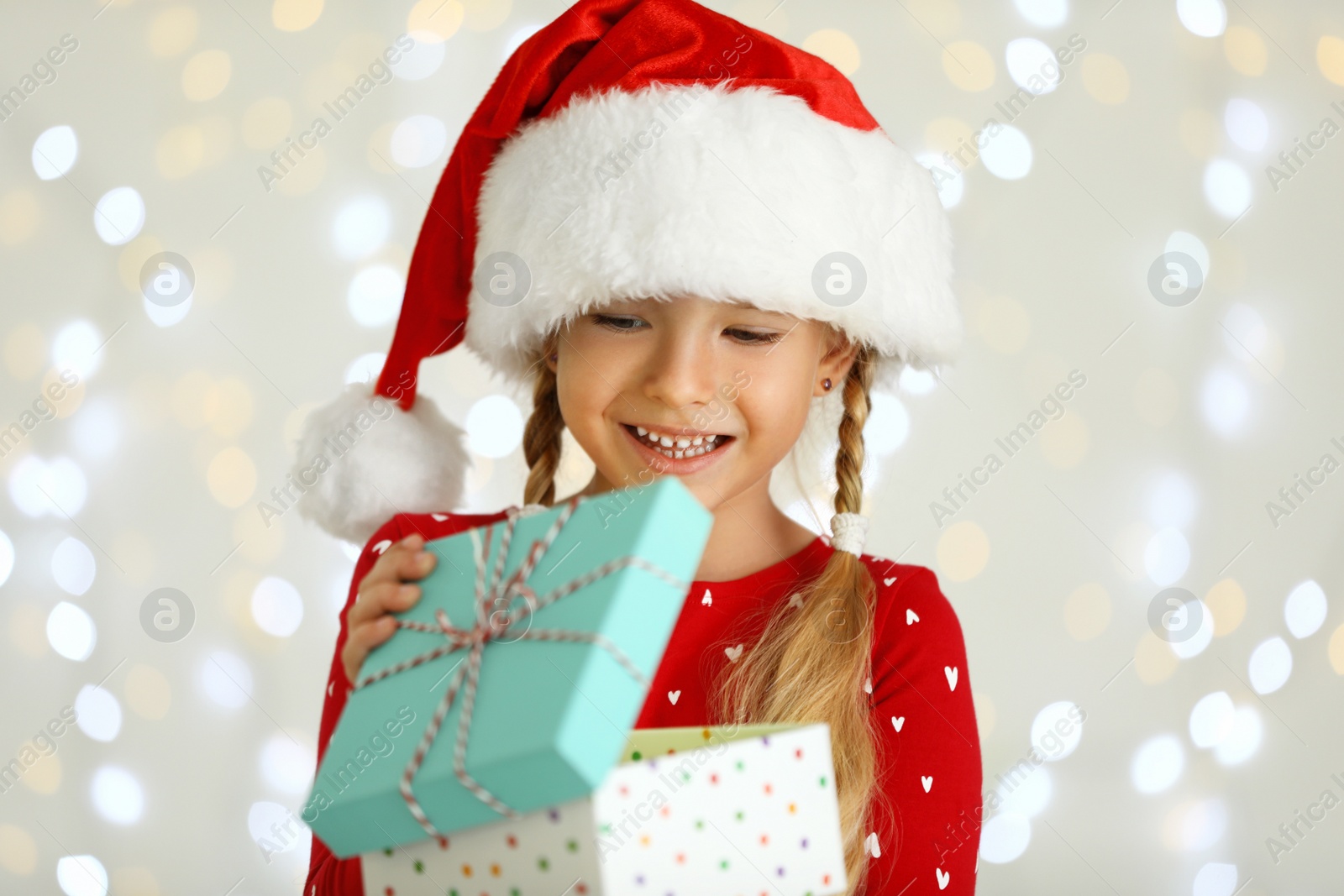 Photo of Happy little child in Santa hat with gift box against blurred festive lights. Christmas celebration