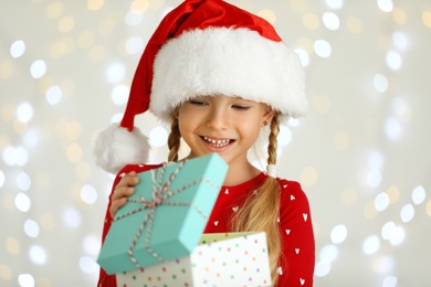 Photo of Happy little child in Santa hat with gift box against blurred festive lights. Christmas celebration