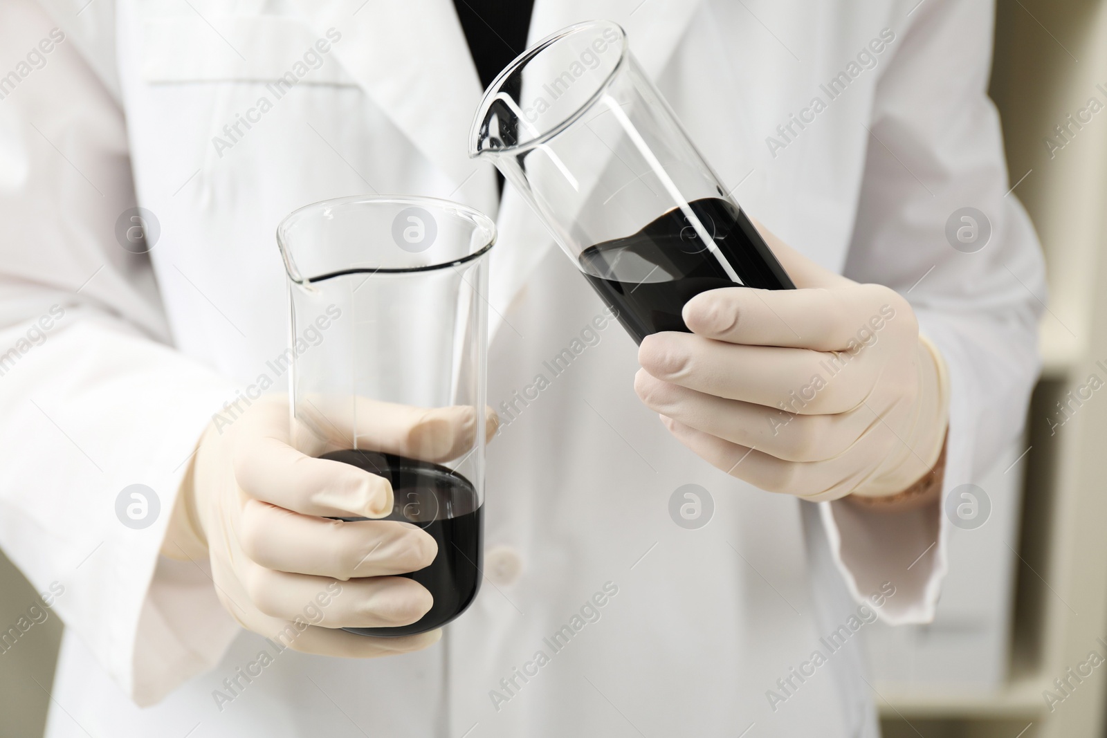 Photo of Laboratory worker pouring black crude oil into beaker indoors, closeup