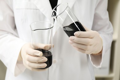 Photo of Laboratory worker pouring black crude oil into beaker indoors, closeup