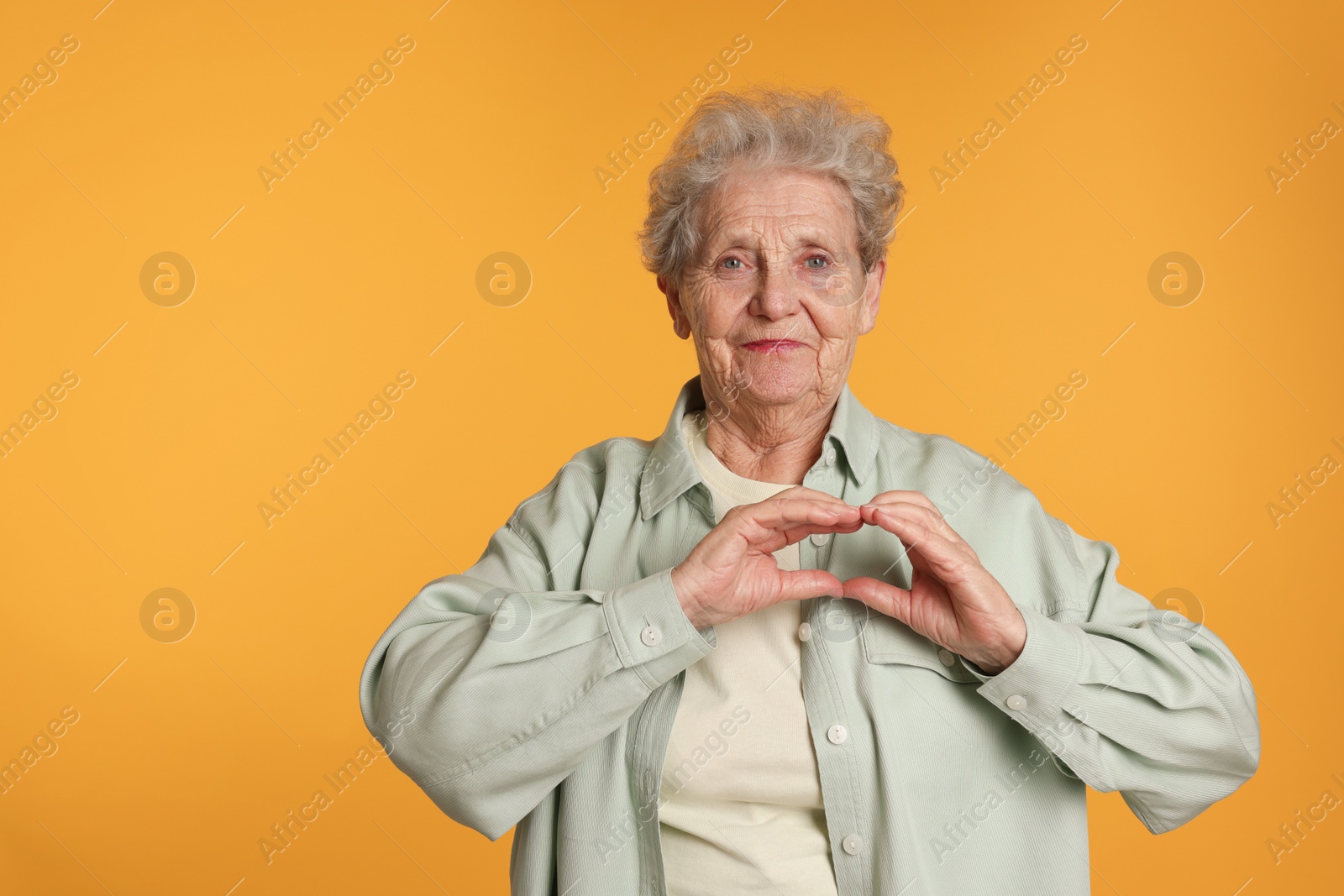 Photo of Elderly woman making heart with her hands on yellow background, space for text