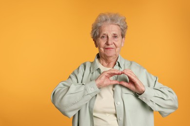 Elderly woman making heart with her hands on yellow background, space for text