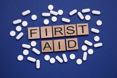 Photo of Words First Aid made of wooden cubes and pills on blue background, flat lay