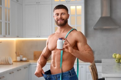 Athletic young man with measuring tape and bottle of supplements in kitchen. Weight loss
