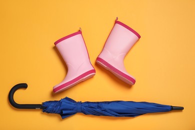 Photo of Pink rubber boots near blue umbrella on orange background, flat lay