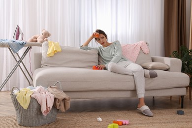 Photo of Tired young mother sitting on sofa in messy living room