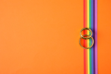 Top view of wedding rings and rainbow ribbon on color background, space for text. Gay symbol