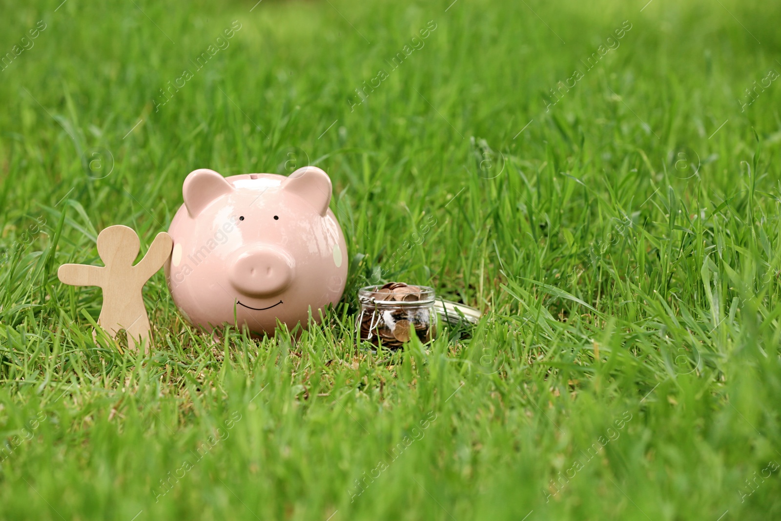 Photo of Piggy bank, wooden person and jar with coins on green grass in park. Space for text