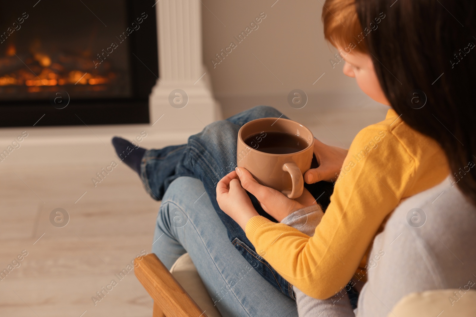 Photo of Mother and son resting together with cup of tea near fireplace at home, closeup