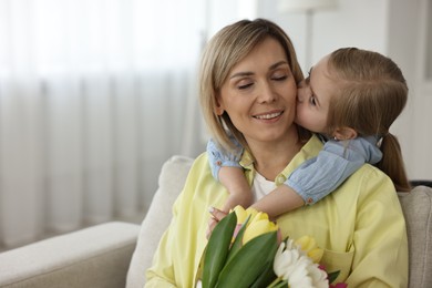 Little daughter kissing and congratulating her mom with Mother`s Day at home, space for text. Woman holding bouquet of beautiful tulips