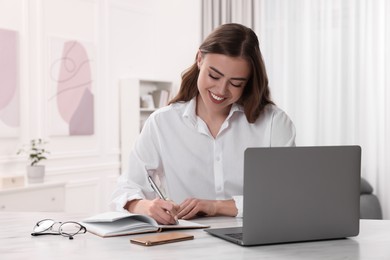 Photo of Happy woman writing something in notebook near laptop at white table in room