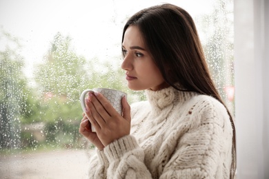 Thoughtful beautiful woman with cup near window indoors on rainy day