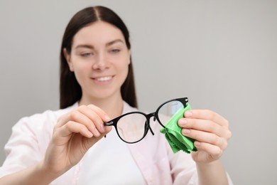 Beautiful woman cleaning glasses with microfiber cloth on light grey background, selective focus