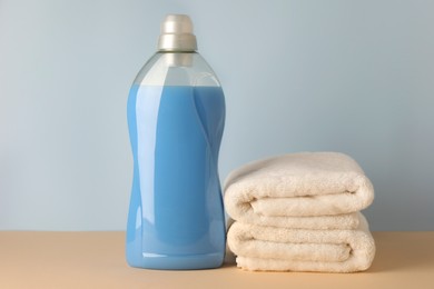 Photo of Bottle of fabric softener and stacked clean towels on pale yellow table against light grey background