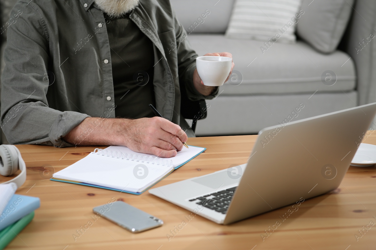 Photo of Man with laptop, notebook and cup of drink learning at wooden table indoors, closeup