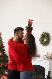 Photo of Happy couple kissing under mistletoe bunch in room decorated for Christmas