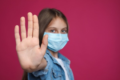 Photo of Little girl in protective mask showing stop gesture on crimson background, space for text. Prevent spreading of coronavirus