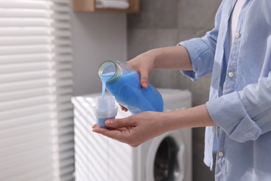 Photo of Woman pouring fabric softener from bottle into cap for washing clothes indoors, closeup