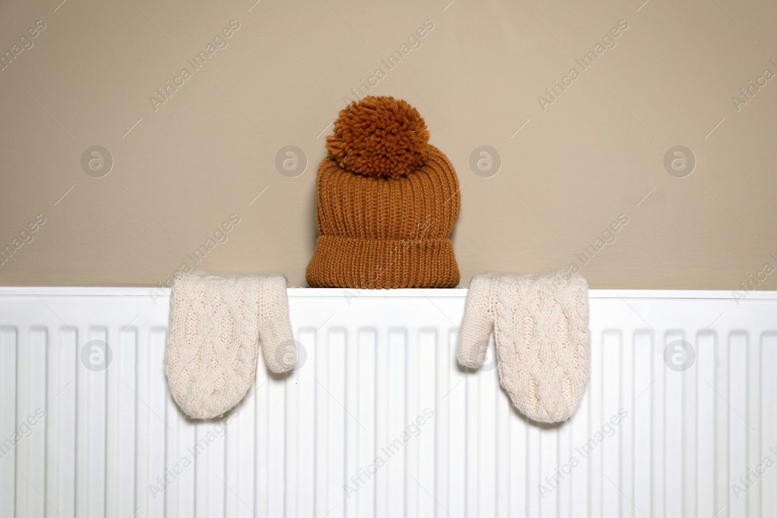 Photo of Modern radiator with knitted hat and mittens near beige wall indoors
