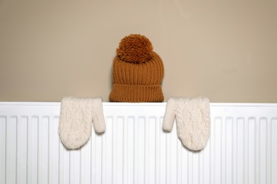 Photo of Modern radiator with knitted hat and mittens near beige wall indoors