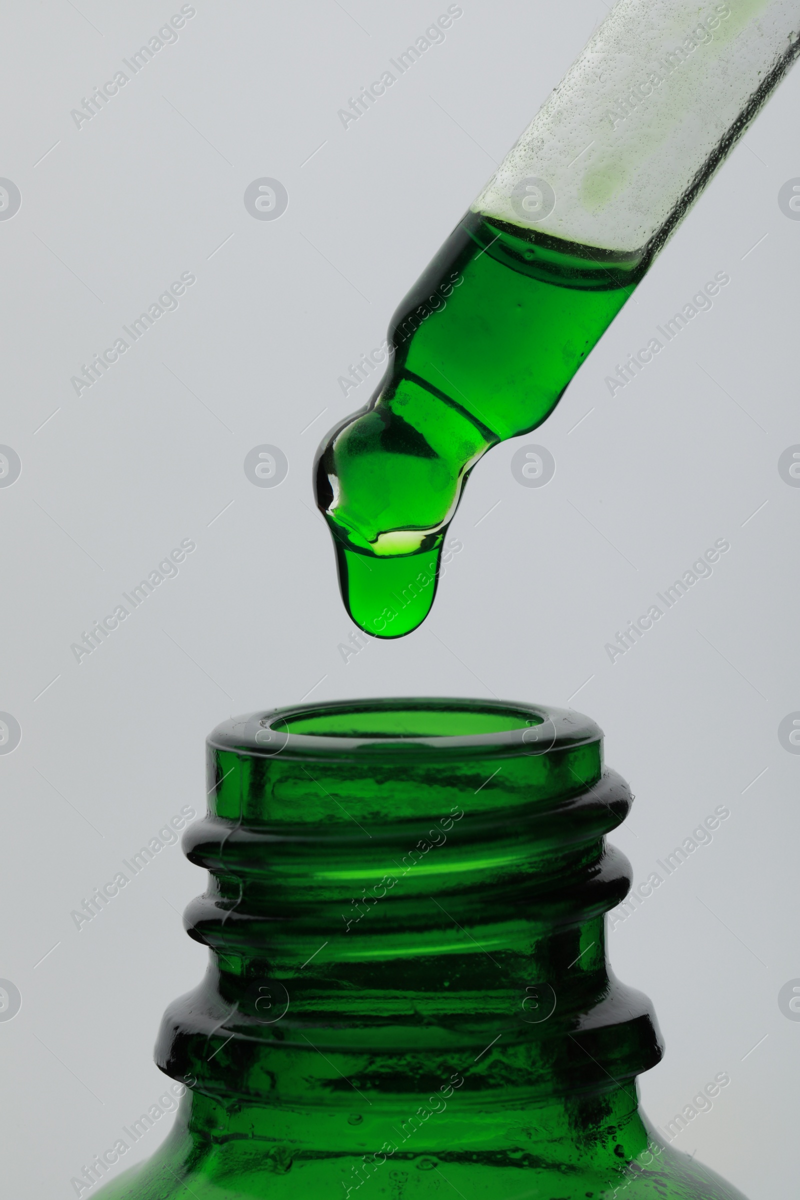 Photo of Dripping green facial serum from pipette into glass bottle on white background, closeup