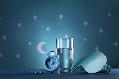 Photo of Alarm clock, soporific pills and sleeping mask near glass of water on table against blue wall decorated with stars and crescent, space for text. Insomnia treatment