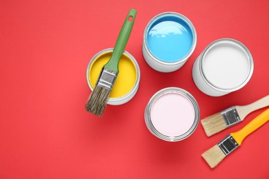 Photo of Cans of colorful paints and brushes on red background, flat lay. Space for text