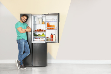 Photo of Happy young man near open refrigerator indoors, space for text